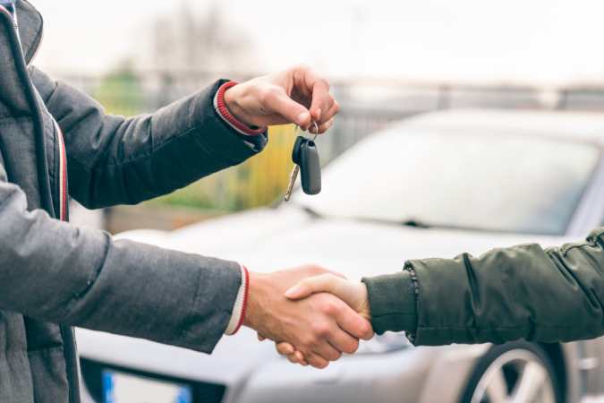 A pair close a sale by shaking hands and exchanging the keys to a new vehicle.